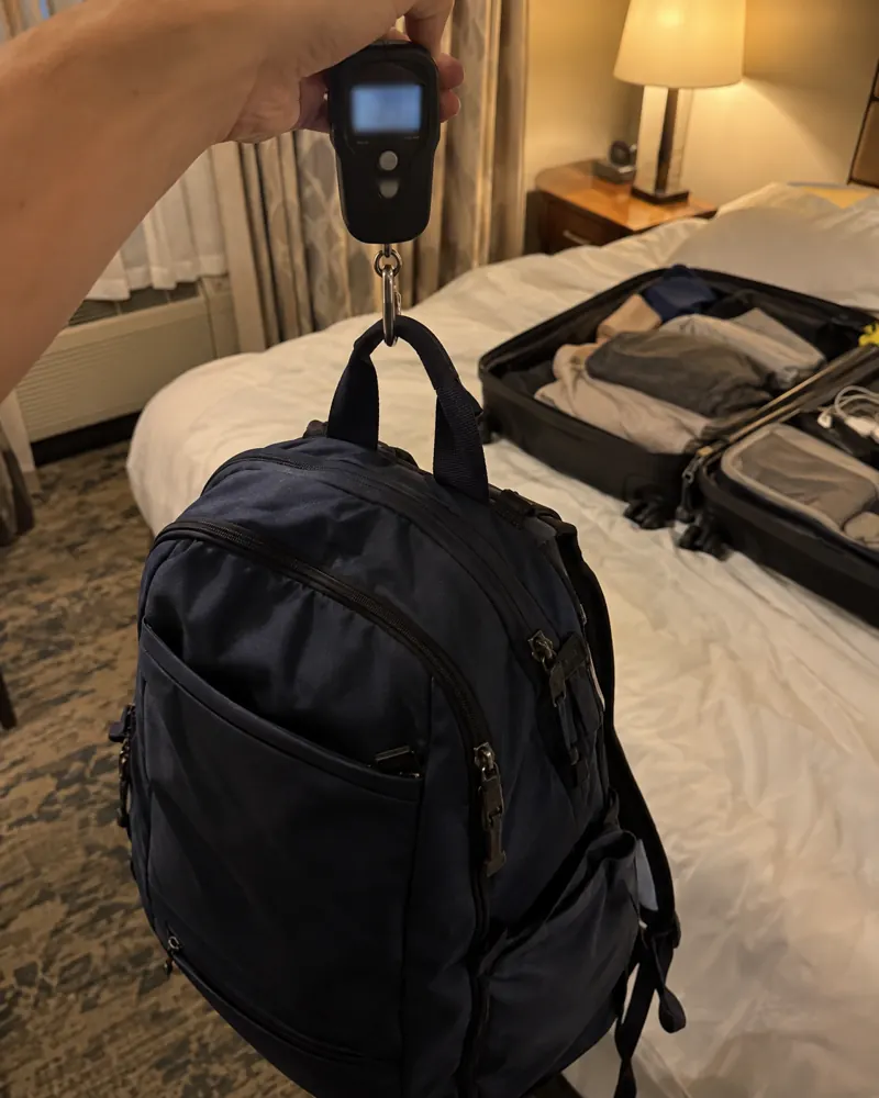 A soft backpack hanging from a handheld luggage scale in a hotel room