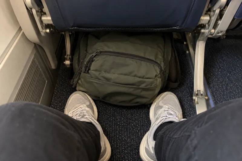 A small backpack stowed under an airplane seat in front of a passenger's feet
