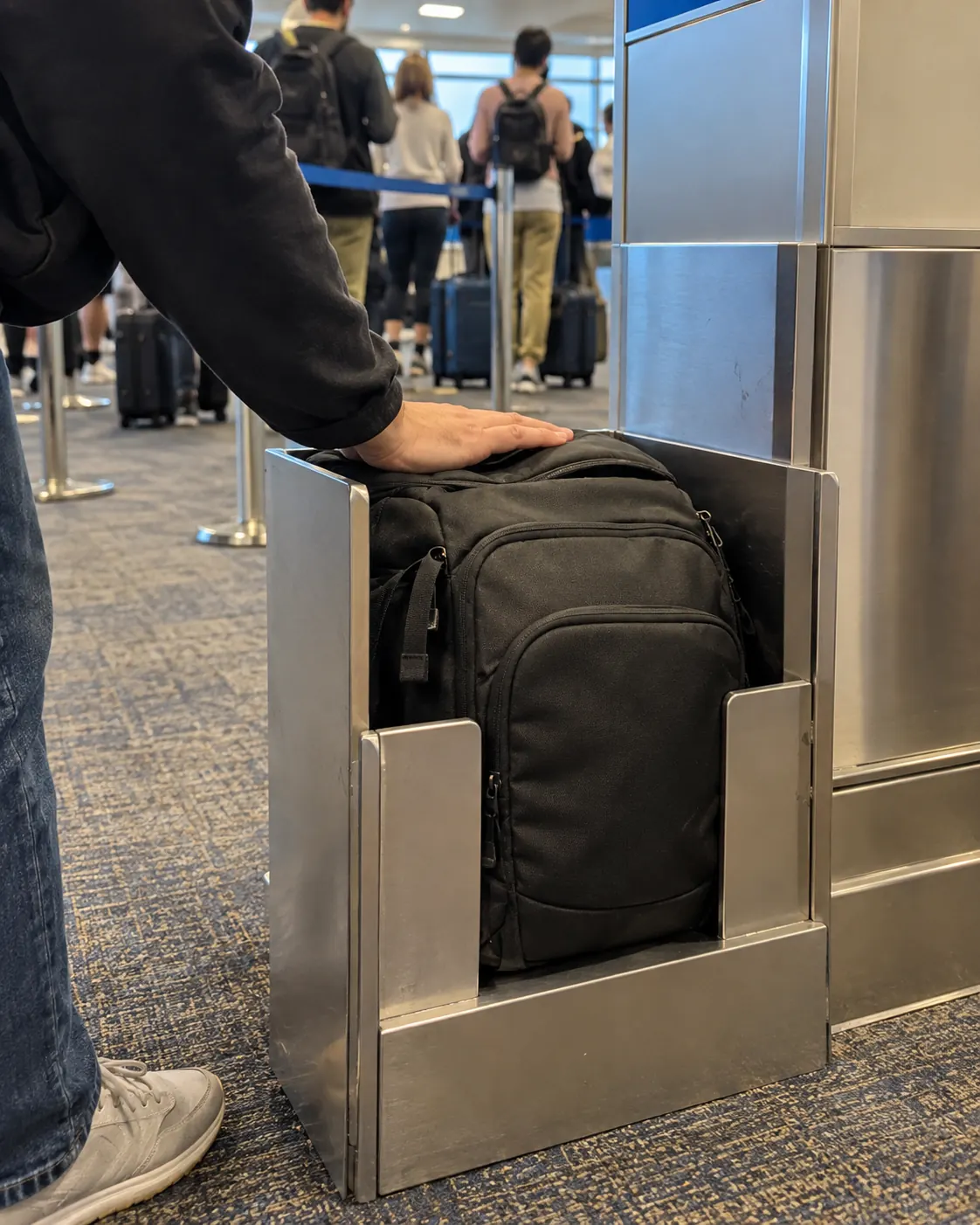 A small black backpack being pushed into a metal bag sizer next to an airport boarding desk