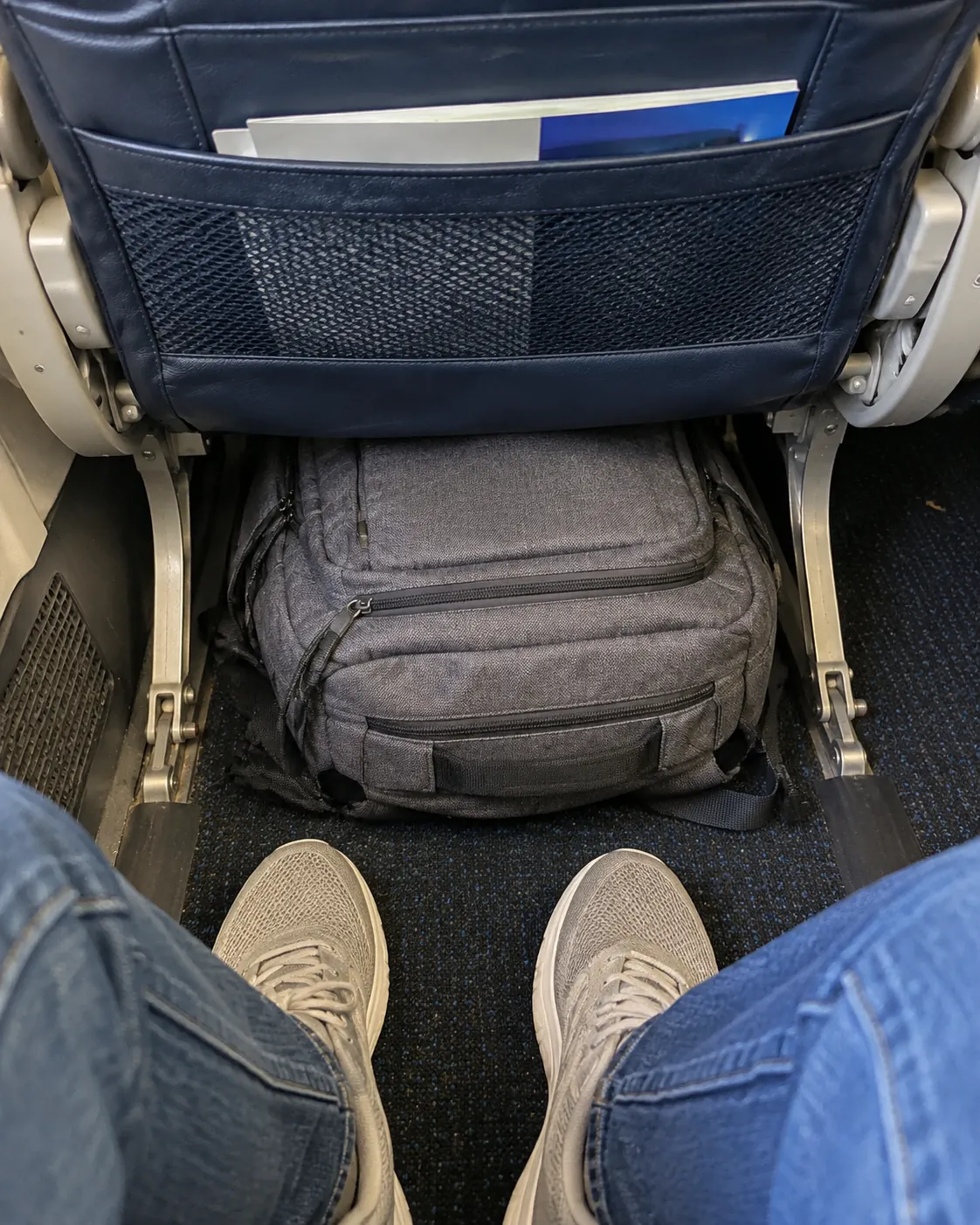 A soft grey backpack pushed under the airplane seat in front, next to a traveler's sneakers
